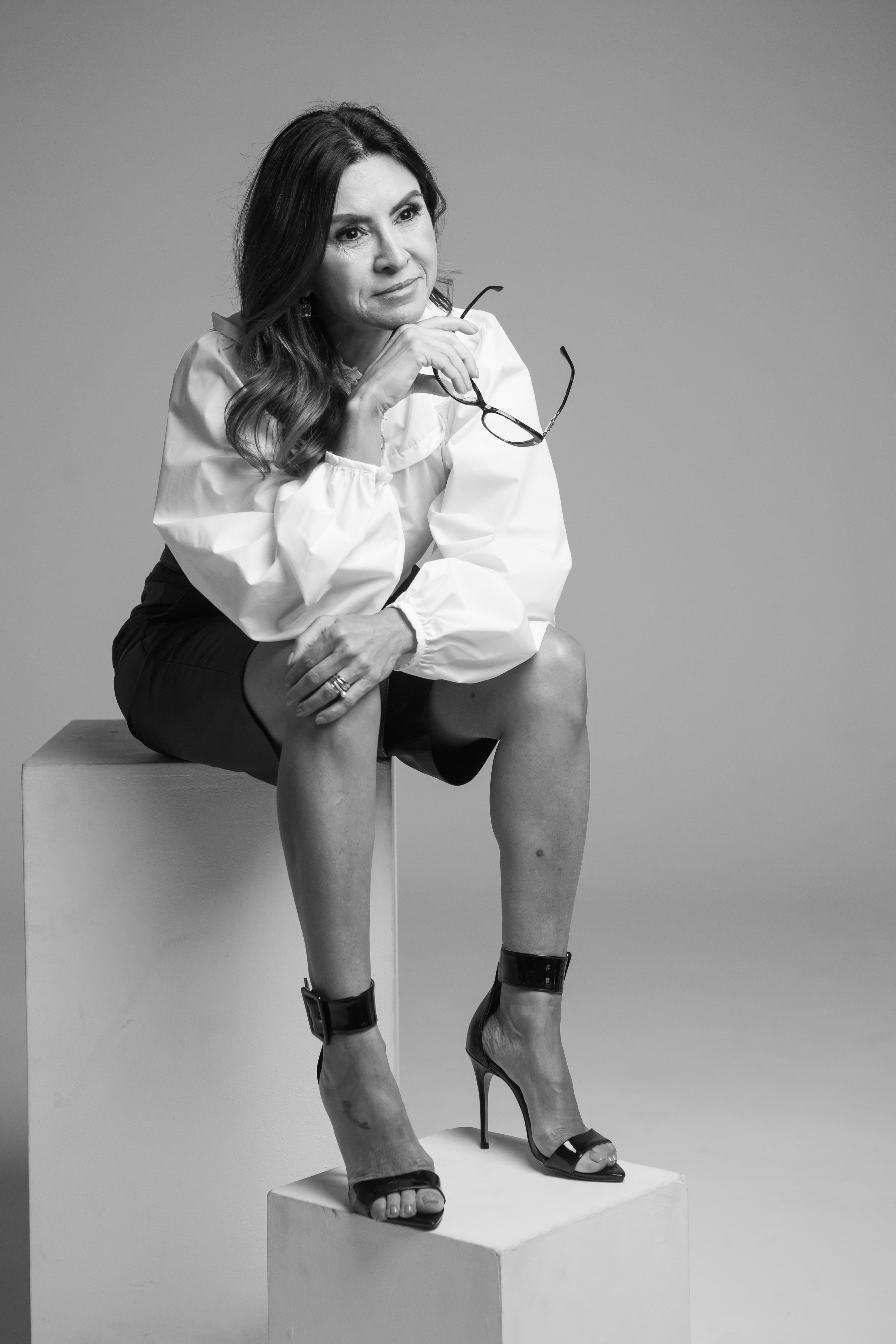 Claudia sentada, óculos na mão, postura premium